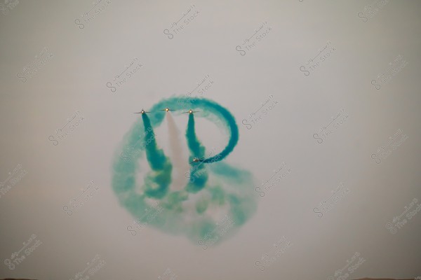 Aircraft perform an exciting aerial display in the sky, leaving behind trails of blue and green smoke, forming circular patterns. The planes fly in precise formation, demonstrating the pilots\' skill, against a pale gray sky.