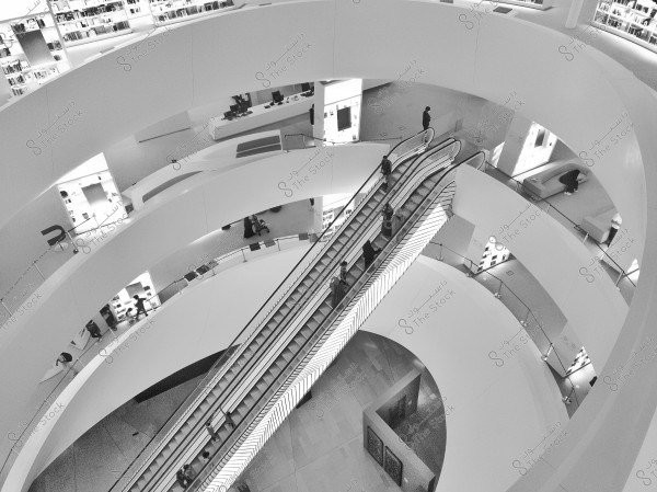 Image of a modern library with a circular design, featuring an escalator in the middle connecting the floors. Books are arranged on shelves surrounding the area, with several people using the escalator and browsing the shelves. The setting is well-lit, creating a calm and organized atmosphere.