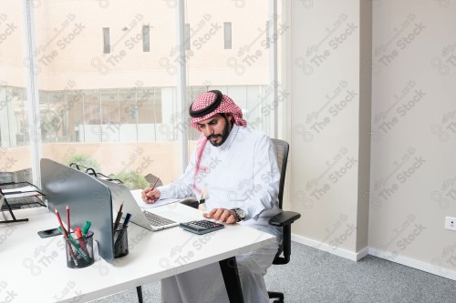 رجل سعودي يرتدي الزي السعودي التقليدي يعمل في مكتب بواجهة زجاجية ويستخدم قلم رصاص لتدوين الملاحظات