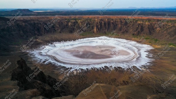 Aerial view of the Al Wahbah crater in Saudi Arabia, showcasing a large crater basin covered with layers of white salt. The crater is surrounded by dark rocky walls, with patches of green vegetation in some areas around it. In the background, barren lands extend to the horizon.