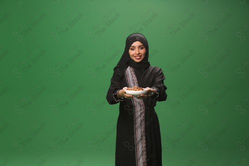 Saudi woman on green background wearing abaya and hijab, making gestures with her hands, presenting a candy tray with a smile, Eid celebrations