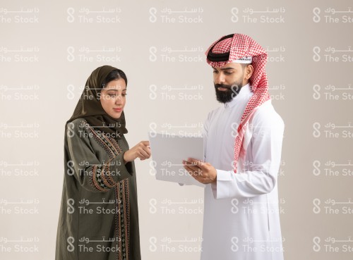 رجل سعودي يرتدي الزي السعودي التقليدي يمسك بالأوراق أثناء حديثه مع امرأة سعودية ترتدي العباءة، زملاء يراجعون بعض المستندات