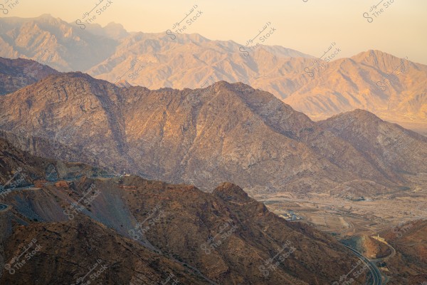 Image of Al Shafa Mountain in Saudi Arabia during sunset, highlighting the rugged rocky terrain of the mountain under the golden sunlight. A winding road is visible, snaking through the mountains, showcasing the natural beauty of the landscape.