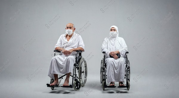 The image shows a man and a woman seated in wheelchairs, both wearing white Ihram garments and face masks. The background is a simple white. This type of clothing suggests they might be participating in Islamic rituals like Hajj or Umrah.