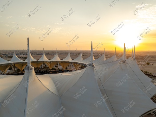 Aerial view of a large fabric-covered building with cone-shaped towers, resembling a sports facility or mall in a Saudi city. The background shows a sunset over the desert and distant buildings.