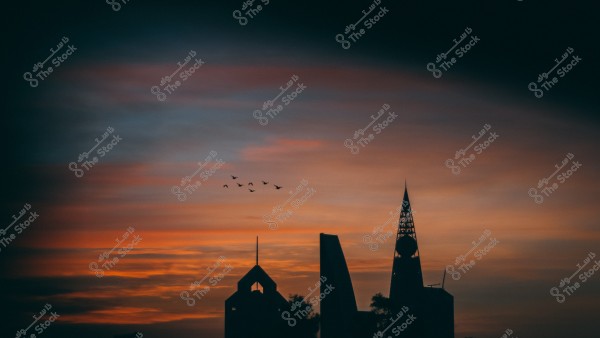A city skyline featuring iconic towers silhouetted against a sunset background. The sky is colored in gradients of orange and purplish-blue, with birds flying in formation across the sky. The buildings have modern designs and appear to be landmarks of the city.