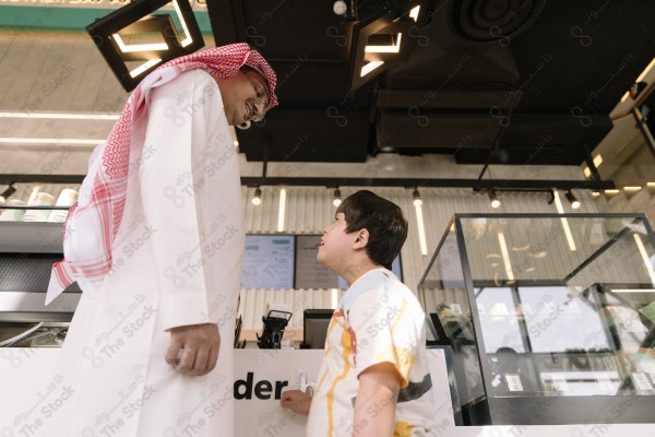 A man wearing a white thobe and red headscarf talking to a child standing in front of an order counter in a cafe.