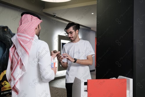 شاب سعودي يرتدي الزي السعودي التقليدي يعرض هاتفه المحمول للموظف ليتأكد من حجزه بينما يحمل بيده أحد المشروبات الغازية