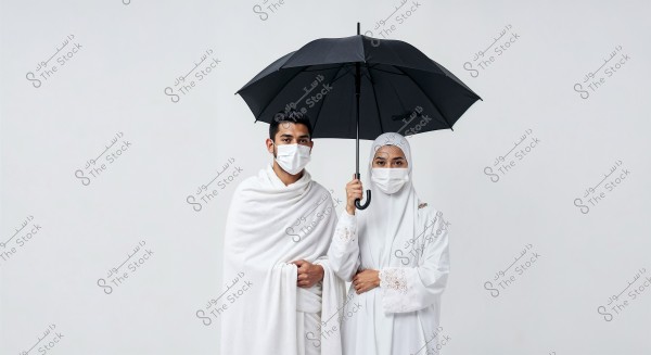 An image of a man and a woman dressed in traditional Hajj attire, with the man wearing a white ihram, and the woman wearing a white long dress and hijab. They stand side by side under a black umbrella in a white studio setting, both wearing medical face masks.