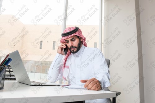 رجل سعودي يرتدي الزي السعودي التقليدي يعمل في مكتب بواجهة زجاجية ويستخدم قلم رصاص لتدوين الملاحظات