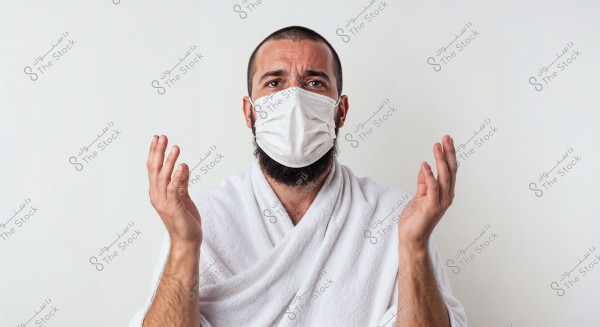 Image of a man wearing a white face mask and a white robe, appearing in a prayer or contemplative pose with his hands raised. The background is plain white.