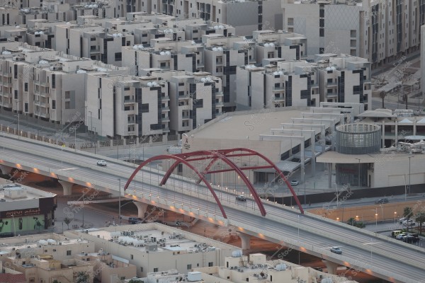 The image depicts an urban scene in a modern city, featuring a collection of residential buildings with contemporary design. A wide bridge with red double arches spans over a highway, adjacent to a large commercial building. The residential buildings are densely arranged throughout the image, with air conditioning units visible on the rooftops. Sparse cars are seen driving on the bridge and highway.