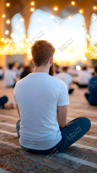A person sitting cross-legged on a large carpet in an open space that appears to be a mosque or plaza, viewed from behind. They are wearing a white shirt and dark-colored pants. The lighting is dim with twinkling lights in the background, and people are seated in rows.