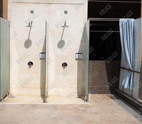 تظهر الصورة مكانًا للاستحمام في الهواء الطلق مع دُشين مزودين برؤوس معدنية كبيرة. يوجد بينهما فواصل زجاجية للتقسيم. الجدران مغلفة ببلاط فاتح اللون. يظهر ستائر زرقاء محيطة بمكان مغلق آخر للاستحمام على اليمين.