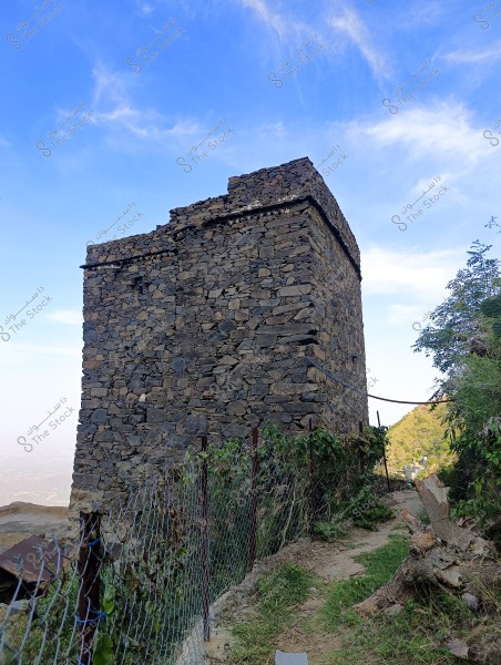 An image of a traditional stone tower located in a mountainous area. The tower is constructed from tightly stacked gray stones, with a metal fence alongside it and surrounded by green vegetation. The sky is clear with a few light clouds, adding to the natural beauty of the scene.