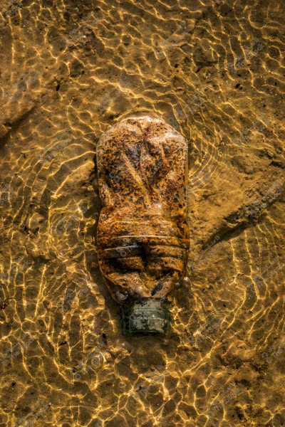 An old, deformed plastic bottle partially submerged in shallow, clear water. The sandy riverbed beneath is visible, with sunlight reflections creating beautiful wavy patterns on the surface.