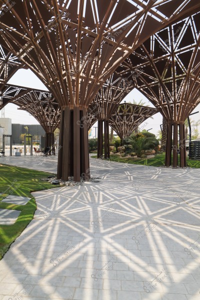 Image of a large metal structure with tree-like branching columns, designed in a unique geometric pattern. Light filters through the spaces, creating beautiful geometric shapes on the tiled floor. In the background, trees and green decorative plants add a natural touch to the scene.