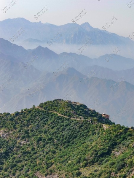 A scenic view of towering mountains covered in lush greenery and dense vegetation, with a small structure or building located at the top of a hill. In the background, a range of connected mountains is visible under a clear sky.