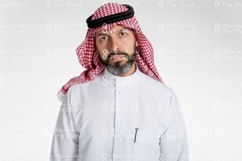 بورتريه رجل سعودي يقف على خلفية بيضاء، يرتدي الثوب، يقف و ينظر بعينيه نحو الكاميرا، ويقوم بعمل ايماءات بوجهه تمثل الجدية او الغضب