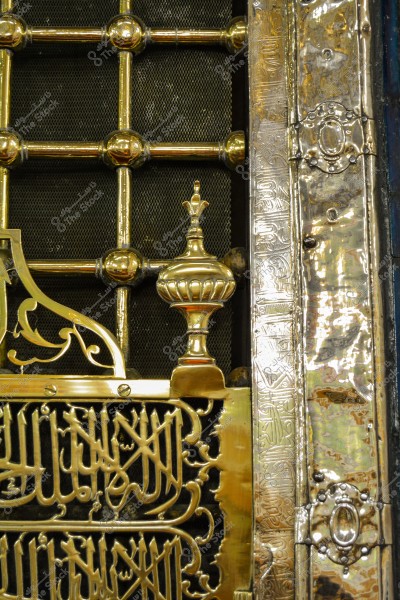 Image of a section of an ornate metal gate featuring intricate Arabic designs with calligraphy. The gate includes golden bars and decorative engravings, representing Islamic architectural art. The image shows detailed decorations and Arabic script etched into the metal.