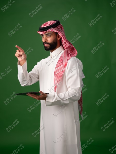 A man wearing a white thobe and a red keffiyeh stands against a green background. He is holding a tablet in his left hand and pointing upwards with his right finger. The clothing suggests he might be from Saudi Arabia.