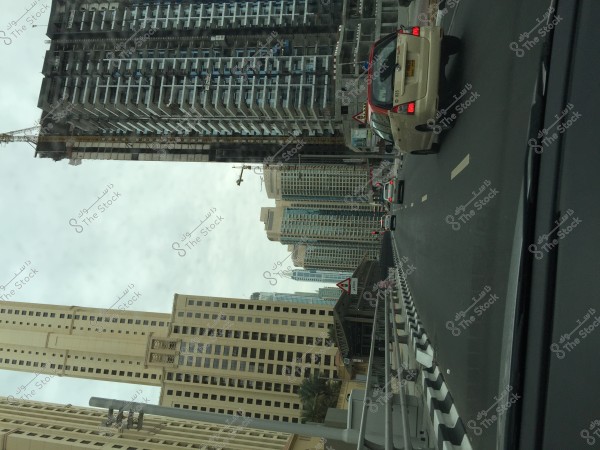 A view of a city street lined with skyscrapers. The image shows several cars driving on a road flanked by tall buildings. The road leads towards a cluster of modern buildings with contemporary architectural design. The weather is cloudy, and the cars observe traffic signs on the road.