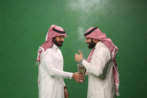 Two Saudi men standing on a green background wearing a thobe and shemagh, interactions and movements of the hands, perfumed with oud and incense, Eid celebrations