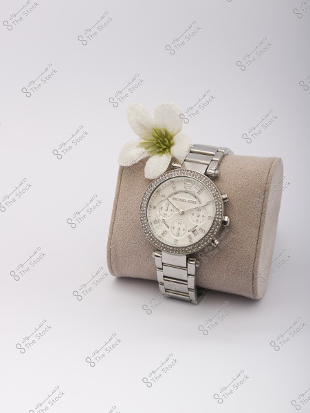 صورة لساعة يد فضية اللون مع حزام معدني, موضوعة على وسادة عرض من القطيفة باللون البيج. تتميز الساعة بوجه دائري محاط بأحجار كريستال لامعة. توجد زهرة بيضاء صغيرة بجانب الساعة, مما يضفي لمسة من الأناقة والرقي على الصورة, وكلها موضوعة على خلفية بيضاء نقية.