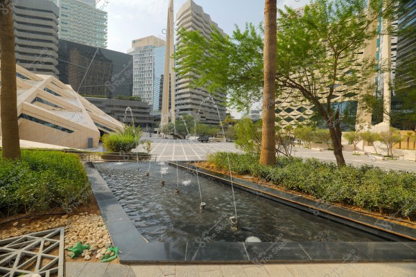 An urban scene showcasing a public square surrounded by modern high-rise buildings, with a small fountain in the center, surrounded by green trees and plants. The area features impressive architecture with innovative angles, clear skies in the background, and wide streets.