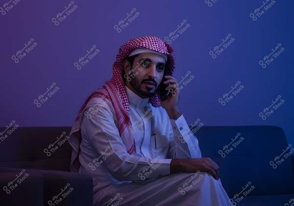 A portrait of a man sitting on a sofa, talking on a mobile phone. He is wearing a white thobe and a red and white checked headscarf, suggesting the attire is from the Gulf region, such as Saudi Arabia. The lighting in the photo features purple and blue hues, creating a modern and calm atmosphere.