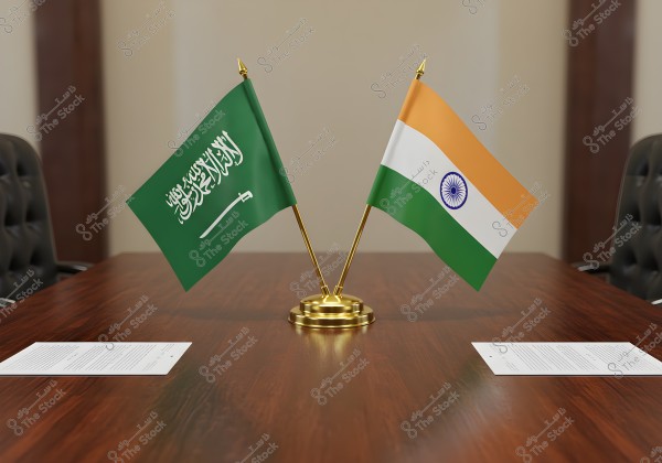 A pair of crossed flags, one of Saudi Arabia and the other of India, mounted on a golden base on a wooden conference table. The image features brown leather chairs on either side of the table, with paper documents placed in front of them.