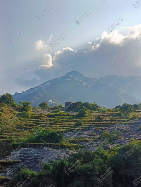 A scenic view showing green agricultural terraces on a mountain slope in sunlight, with dense clouds covering the mountain peak and rays of sunlight breaking through. There are ruins of an old structure amidst the greenery, while some buildings are visible in the distance at the top of the mountain under the sunny sky.