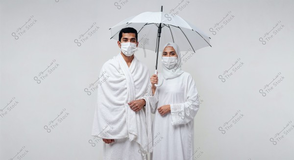 An image of a man and woman wearing traditional white ihram clothing. The man is dressed in two white wraps, and the woman is wearing a hijab and a white dress while holding a white umbrella. Both are wearing medical masks. The image appears to be related to the rituals of Hajj or Umrah.