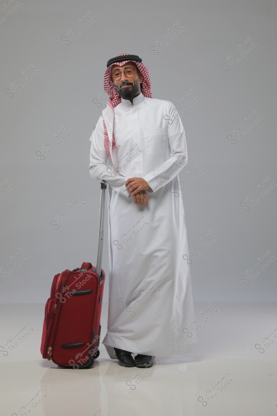 An image of a man wearing a Saudi thobe with a red ghutra and agal, standing next to a red suitcase on a white floor with a gray background.