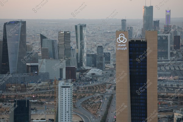 Aerial view of Riyadh city, showcasing a collection of modern skyscrapers and buildings. In the foreground, a tall tower has the logo and name \"Al Rajhi Bank\" illuminated on the side. The image features wide roads and diverse architectural structures filling the city below, with the horizon visible in the background under a cloudy sky.