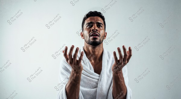 A portrait of a man wearing a white robe, raising his hands towards the sky with an intense expression on his face. He is shown from the upper body with a light-colored background, highlighting his facial expression and open hands.