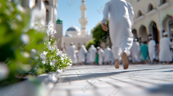 The image shows a paved street with a group of people wearing white thobes walking towards a mosque, featuring a minaret and dome in the background. On the left side of the image, there are white and green flowers along the path.