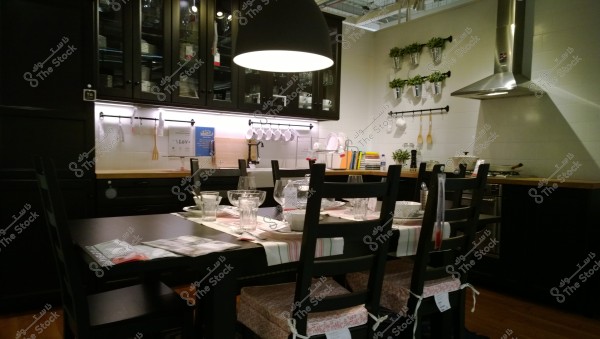An image of a modern kitchen interior with a stylish design featuring a black dining table with chairs padded with floral patterns. The table is set with glassware and dishes on red-striped fabric napkins. In the background, black cabinets with a wooden countertop and hanging kitchen tools on a white tiled wall can be seen. There is a metal vent hood above a stove and herb plants decorating the walls.