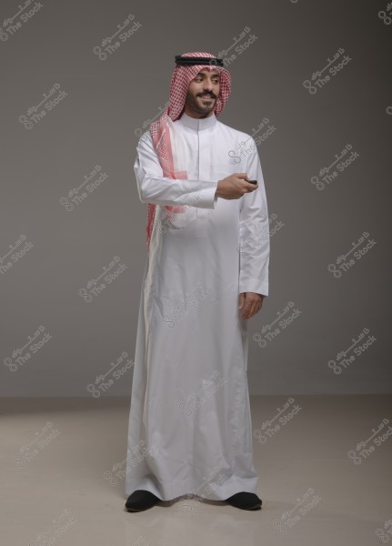 صورة لرجل يقف في استوديو يرتدي ثوبًا أبيض وغترة مع عقال, وهو زي تقليدي شائع في السعودية. يبدو أنه يشير بيده اليُمنى. الخلفية محايدة ذات لون رمادي.