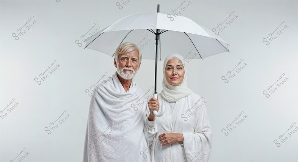 A photo of a man and woman standing side by side under a white umbrella. The man is wearing traditional white Hajj attire, and the woman is wearing a white hijab and abaya, suggesting they are ready for performing Hajj rituals. The background is plain white with no additional details.