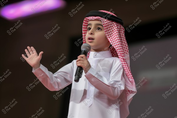 A portrait of a child wearing traditional Gulf attire, including a white thobe, a red and white checkered headscarf (ghutra) with a black agal. The child is speaking into a microphone, with one hand raised, suggesting he is giving a speech or participating in an event.