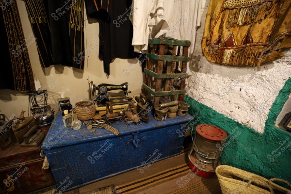 An image depicting a collection of traditional tools and items inside a room. There is a blue chest at the forefront topped with an antique sewing machine, alongside various objects like wicker baskets and hand grinders. Traditional embroidered garments are hung on the walls, reflecting rich heritage. A lantern, a kettle, and an old heater are also visible on the right side of the image.