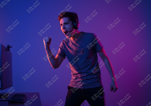 Image of a young man wearing a headset, playing on a computer. The background is illuminated in blue and purple tones. The young man is wearing a gray t-shirt and track pants, appearing excited as if celebrating a win or success moment in the game.