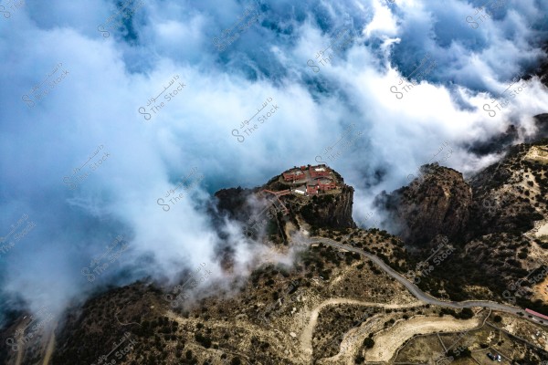 An aerial view showing a castle or a collection of buildings atop a mountainous peak surrounded by clouds. Winding mountain roads and rugged terrain surround the site. The dense clouds create a dramatic scene, giving the image a sense of mystery and intrigue.