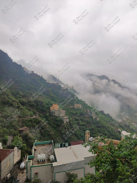 The image depicts a natural landscape of mountains covered in greenery in a mountainous area, with buildings scattered along the mountain slopes. Low clouds permeate the scene, enhancing the natural beauty of the location. The image also shows an overcast sky, evoking a sense of calm and tranquility.