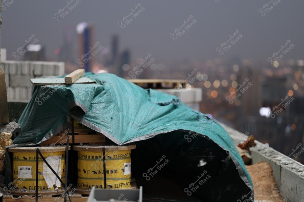 A scene of a construction site at night where a green fabric cover is draped over construction materials placed on the rooftop of a building under development. In the background, the blurred city lights illustrate tall buildings with glowing lights against a grey sky.