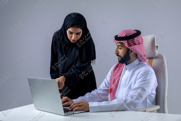 A photo of an Arab man and woman in a work setting. The man is seated on a chair wearing a Saudi thobe and red shemagh, working on a laptop. The woman stands beside him, pointing at the computer screen, wearing a black abaya and hijab. Both appear focused and engaged in their work. The background is a light grey neutral color.