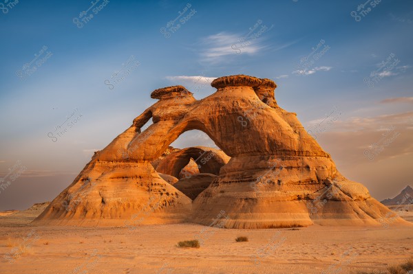 An image of a rock formation in the desert featuring a natural arch made of reddish sandstone. The formation has a rough surface and massive dimensions, with a blue sky and scattered clouds in the background. The scene appears to be set in a desert area possibly in Saudi Arabia.