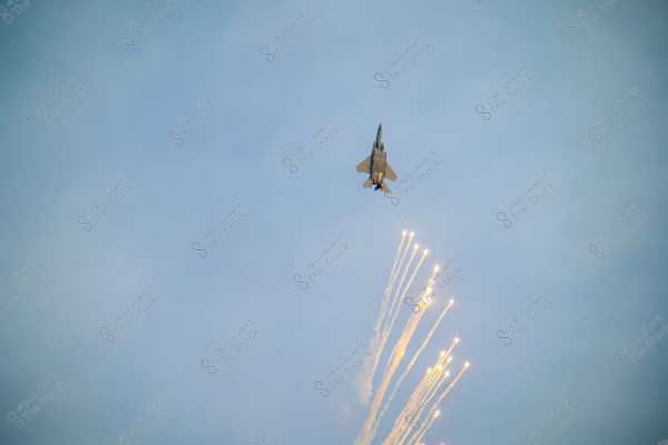 The image shows a fighter jet flying upward in the sky while releasing bright flares. The background is a clear blue sky, and the bright flares create a dramatic effect with their vivid glow.