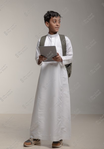 A portrait of a boy standing in the center of the frame, wearing a white thobe and a gray backpack. He is holding a tablet and looking to the side. He is wearing sneakers and is in an indoor setting with soft lighting, conveying a sense of attentiveness and preparedness.